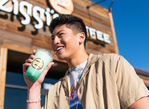 A smiling man holding a Ziggi's Coffee green iced drink outside a Ziggi's Coffee location on a sunny day.