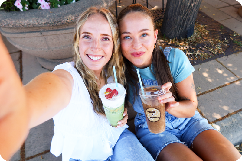 Two smiling women taking a selfie outdoors, holding Ziggi's Coffee drinks including an iced matcha with strawberry topping and an iced coffee.