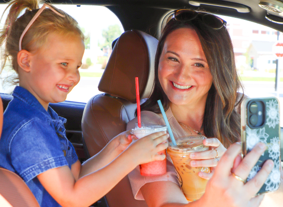 A smiling woman and young girl sitting in a car taking a selfie, each holding a Ziggi's Coffee drink including a pink iced drink and an iced coffee.