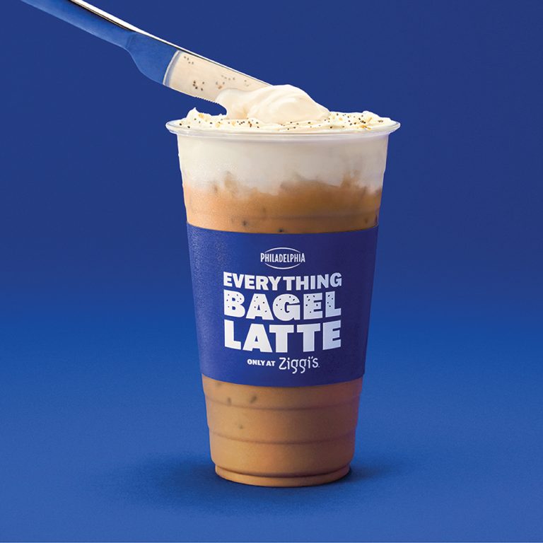 Philadelphia × Ziggi's Coffee Everything Bagel Latte on a blue background — an iced latte topped with cream cheese cold foam and everything bagel seasoning, being spread with a knife.