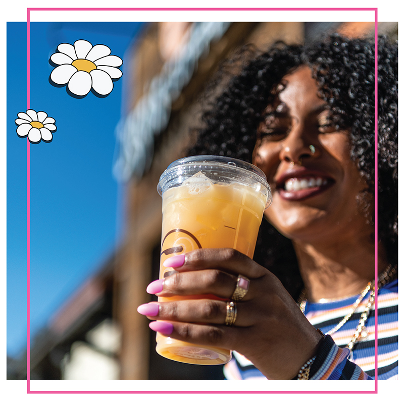 A smiling woman with curly hair holds a Ziggi's Coffee branded iced orange drink outdoors under a blue sky, with daisy illustrations overlaid on a pink-bordered photo.