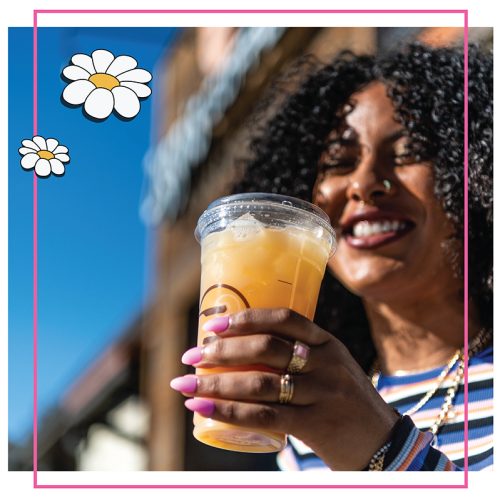 A smiling woman with curly hair holds a Ziggi's Coffee branded iced orange drink outdoors under a blue sky, with daisy illustrations overlaid on a pink-bordered photo.